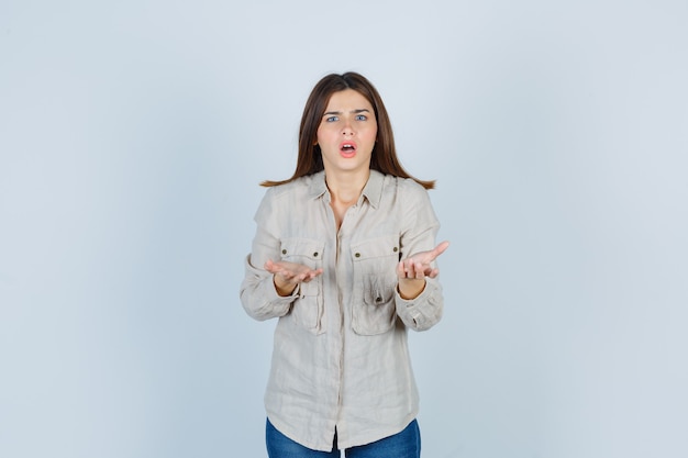 Young girl stretching hands in questioning manner in beige shirt, jeans and looking baffled , front view.