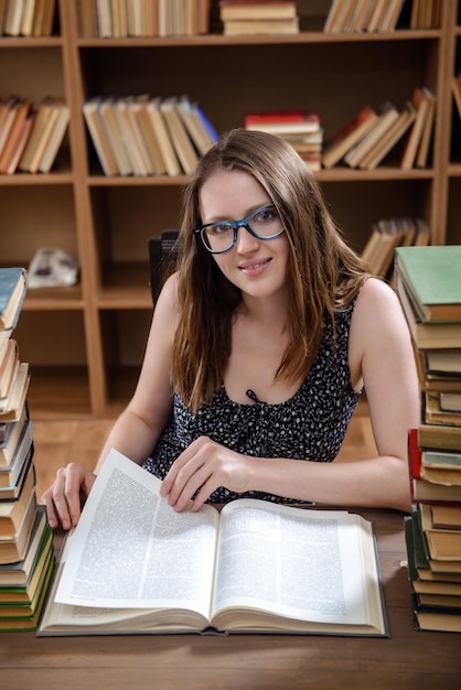 Young girl student reads a textbook sitting at a table with many books in the library