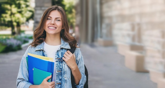 Young girl student smiling against university holds folders and notebooks in hands