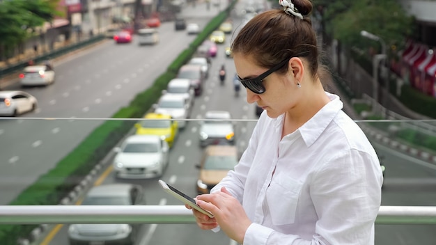 Young girl in sunglasses on modern smartphone standing on bridge on the background traffic jam close up