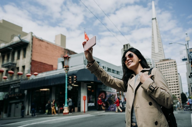 Photo young girl taking selfie standing on road in chinatown. female asian travel backpacker holding cellphone make self portrait with transamerica pyramid tower building in san francisco in usa.