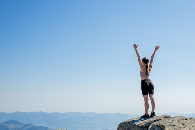 The young girl at the top of the mountain raised her hands up on blue sky. The woman climbed to the top and enjoyed her success. Back view.