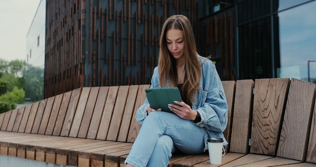 Young girl using tablet computer outdoors in the street while sitting on the bench Pretty female tourist strolling and looking at screen browsing Internet chatting