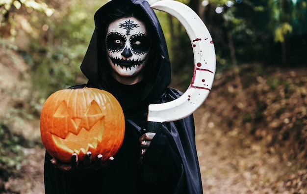 Young girl wearing mysterious Halloween dress holding scary pumpkin and sickle with blood in the woods - Focus on face