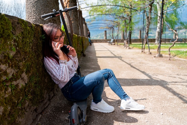 Young girl wearing a shirt and headphones and talking on the phone riding a scooter