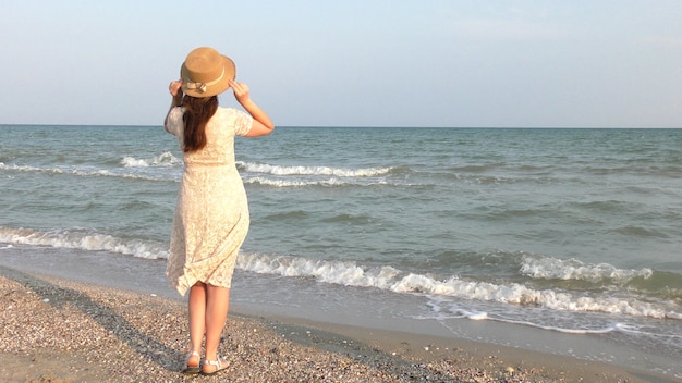 Young girl wearing white lace dress and hat standing on tropical beach. Blue sea on the background. Summer vacation concept