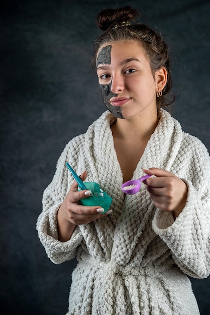 Young girl in white bathrobe applying restorative black mask on face isolated on dark background