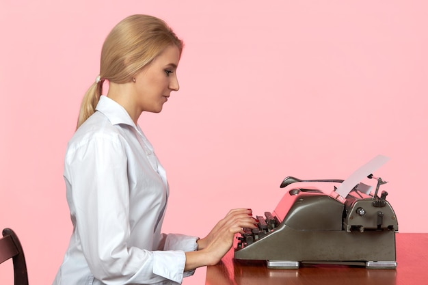 A young girl in a white blouse sits at a desk in the office and types text on a retro typewriter.