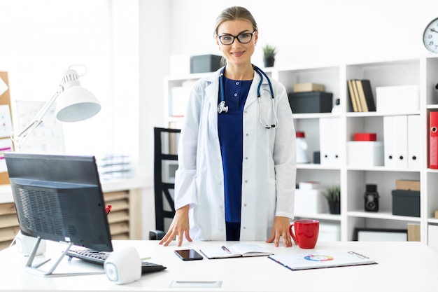 A young girl in a white coat is standing near a table in her office. A stethoscope hangs around her neck.