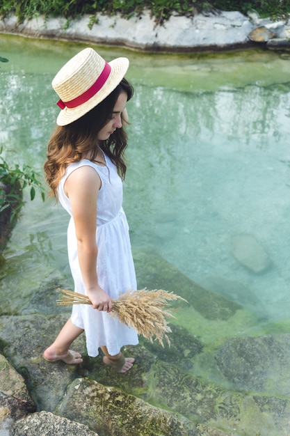 A young girl in a white dress on a sunny day enters the lake. Girl in a straw hat in the river at sunset.