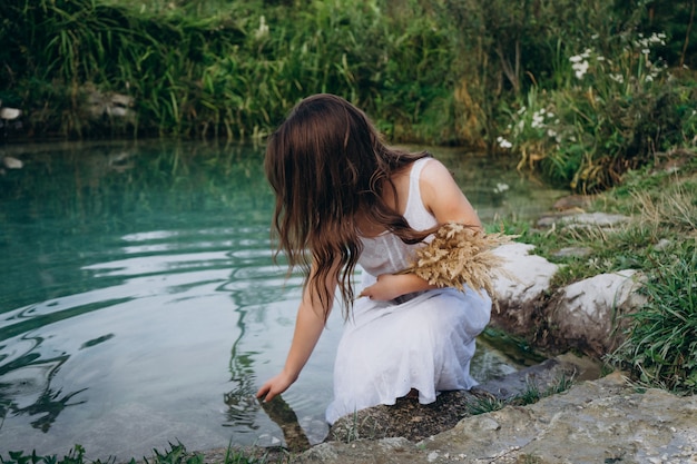 Young girl in a white dress on a sunny day near the lake in the woods. Girl in a straw hat on the shores of a forest lake. Beautiful blue lake among the forest.