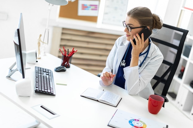 A young girl in a white robe is sitting at the desk in the office, talking on the phone and holding a pen