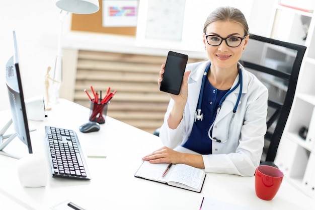 A young girl in a white robe sits at a table in the office and holds a phone in her hand. A stethoscope hangs around her neck.