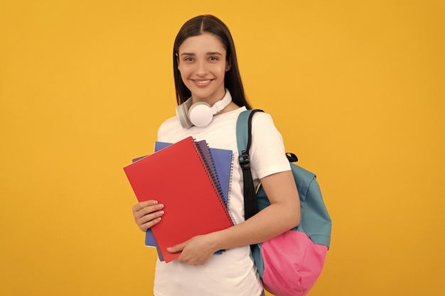 Young girl with backpack and headphones hold workbook on yellow background back to school