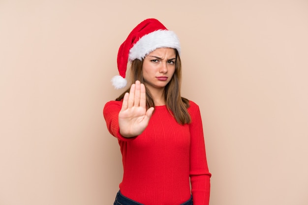 Young girl with christmas hat over isolated wall making stop gesture with her hand