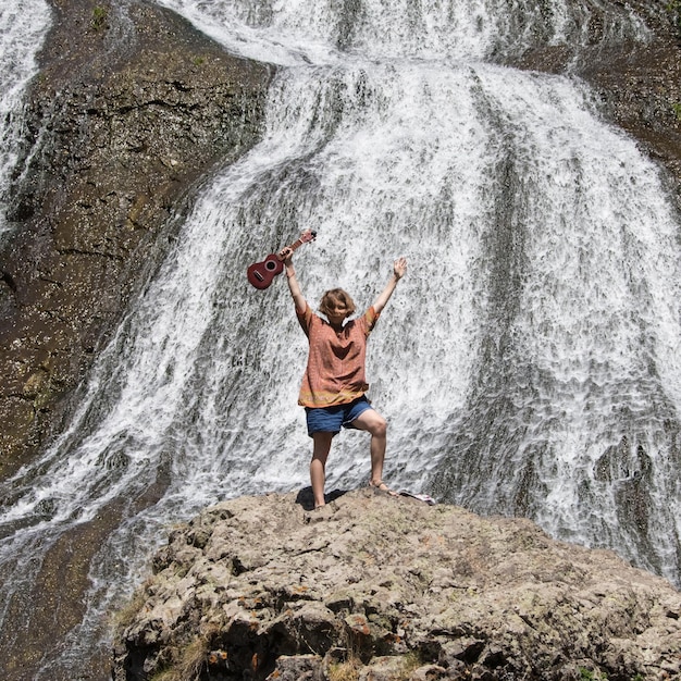 Young girl with hands up with ukulele standing on a rock near waterfall