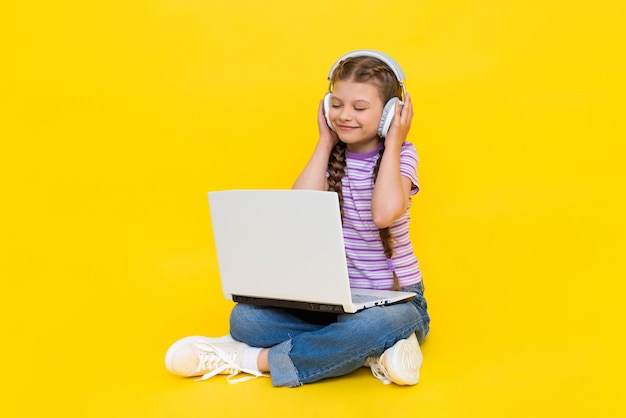 A young girl with a laptop A little girl is sitting crosslegged on the floor holding a laptop in her hands and listening to music with headphones Music for teenagers Yellow isolated background