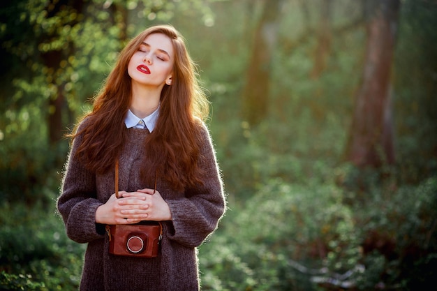 A young girl with long hair in a warm sweater in the forest holds an old film camera in her hands