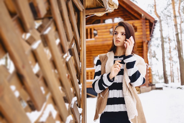 Young girl with phone in waistcoat on against of wooden cottage in winter forest