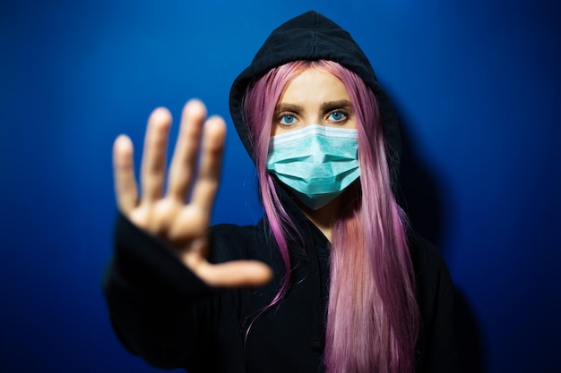 Young girl with pink hair and blue eyes, wearing medical flu mask and hooded sweater, showing Stop gesture on wall of phantom blue color.