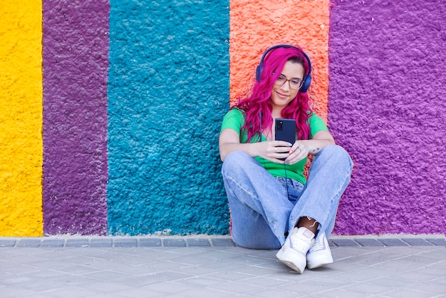 Young girl with pink hair sitting on the street with colorful background and listening to music wit