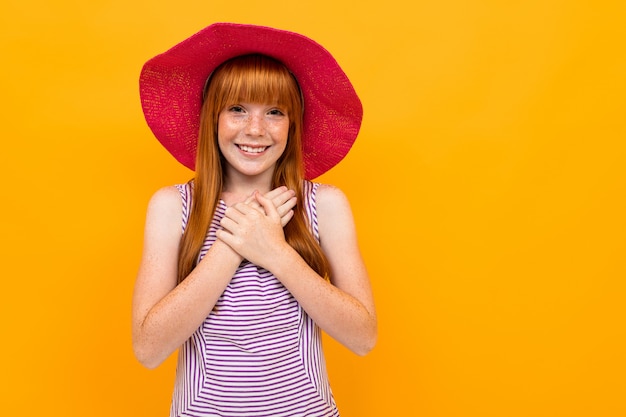 Young girl with red hair and pink hat smiles and hopes for something isolated on yellow