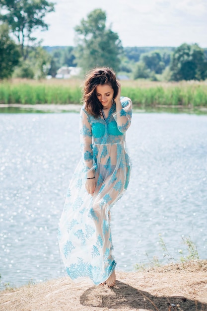 Young girl with a sexy body in a swimsuit and tunic smiles on beach by the lake