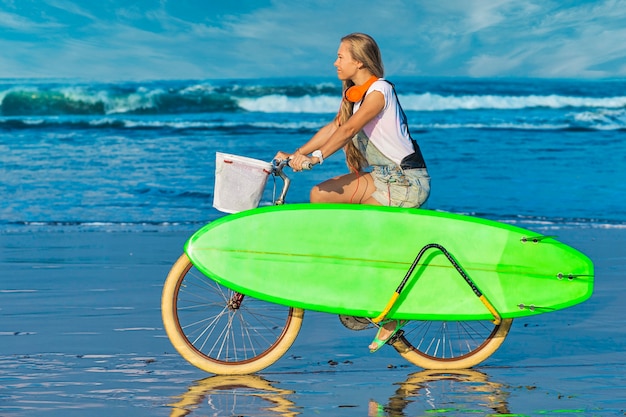 Young girl with surfboard and bicycle on the beach 