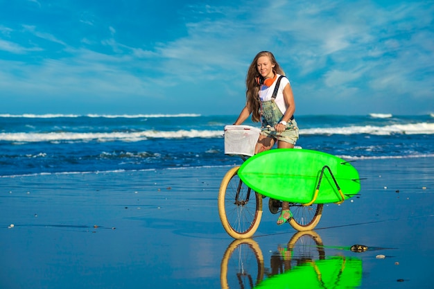 Young girl with surfboard and bicycle on the beach 