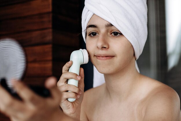 Young girl with towel on her head makes facial massage with wireless electric massager