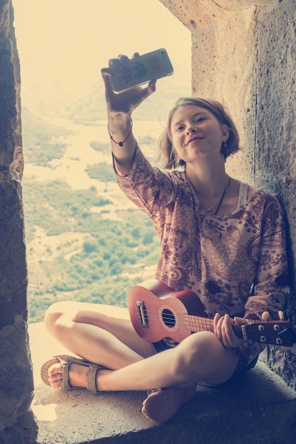 Young girl with ukulele sitting in arched window and taking photo selfie