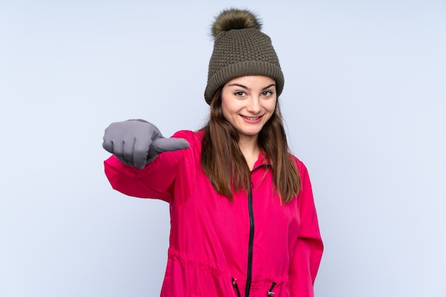 Young girl with winter hat isolated on blue wall giving a thumbs up gesture