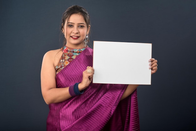 A young girl or woman wearing an Indian traditional saree holding a signboard in her hands on gray background