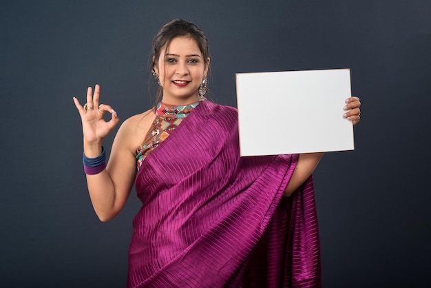 A young girl or woman wearing an Indian traditional saree holding a signboard in her hands on gray background