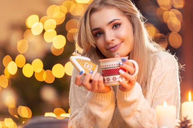 A young girl in a wool sweater holding a gingerbread and a cup with tea coffee or Christmas punch