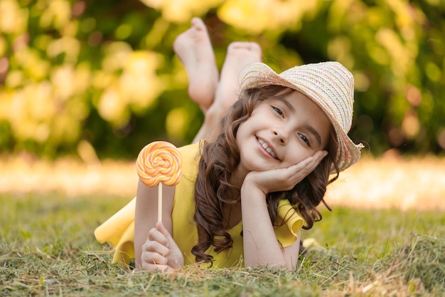 A young girl in a yellow suit and a hat with a lollipop in her hands outdoors in the park