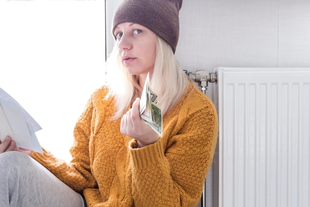 A young girl in a yellow sweater and a brown hat is sitting on the floor counting money and thinking how to pay bills and taxes near a heater with a thermostat