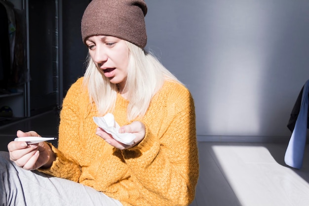 A young girl in a yellow sweater and a brown hat looks at the thermometer in surprise which shows a high temperature