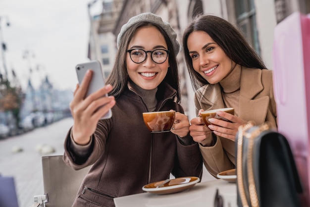 Young girls friends sitting outdoors in cafe drinking coffee and taking selfie on smartphone