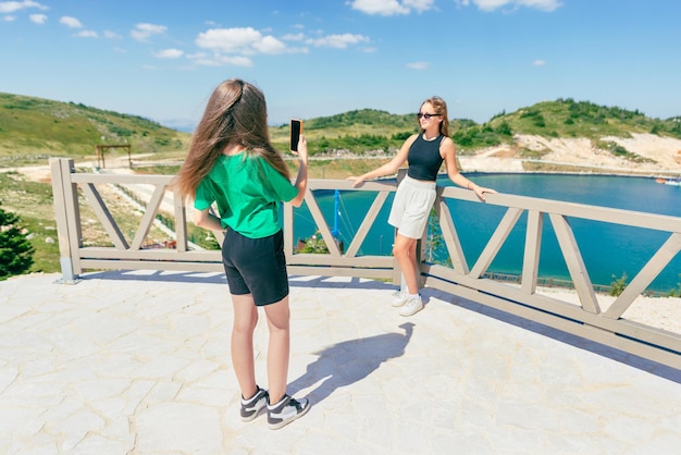 Young girls taking photos with phone of each other Tourist teenage girls on the mountain by the lake