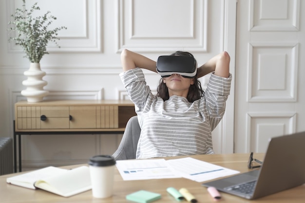 Young glad woman office worker in virtual reality glasses at workplace relax in VR helmet at work