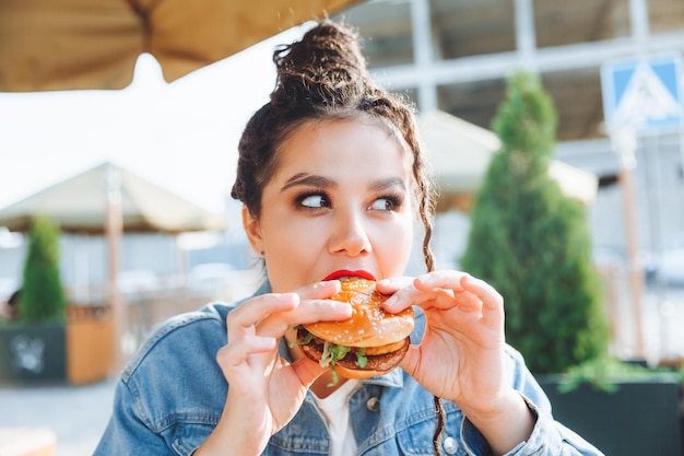 A young glamorous woman with dreadlocks and red lipstick is sitting and eating a burger in a street cafe the concept of eating longlasting lipstick