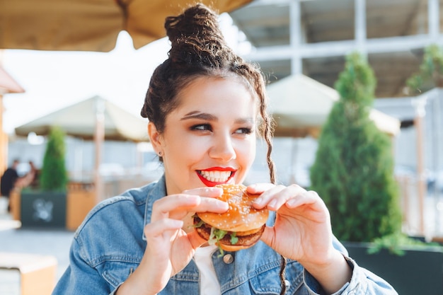 A young glamorous woman with dreadlocks and red lipstick is sitting and eating a burger in a street cafe the concept of eating longlasting lipstick