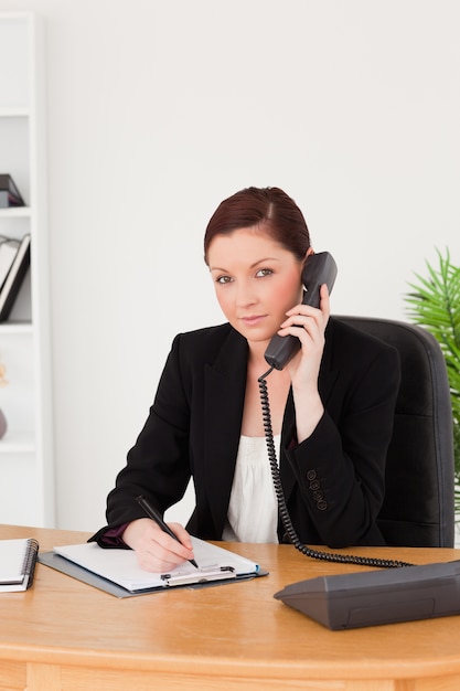 Young gorgeous red-haired woman in suit writing on a notepad and phoning