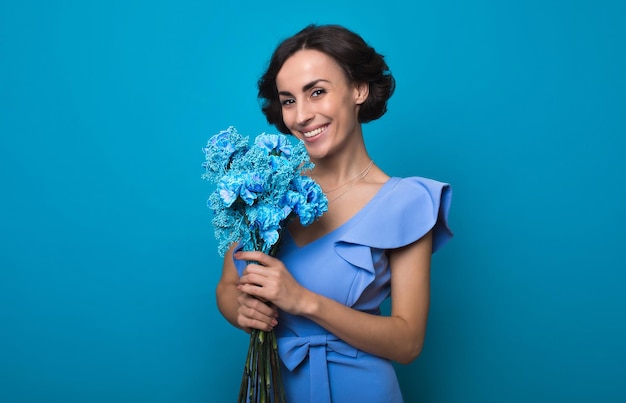 Young gorgeous woman in a bright blue dress is looking in the camera with a big smile holding a bunch of blue flowers in her hands International Women's Day