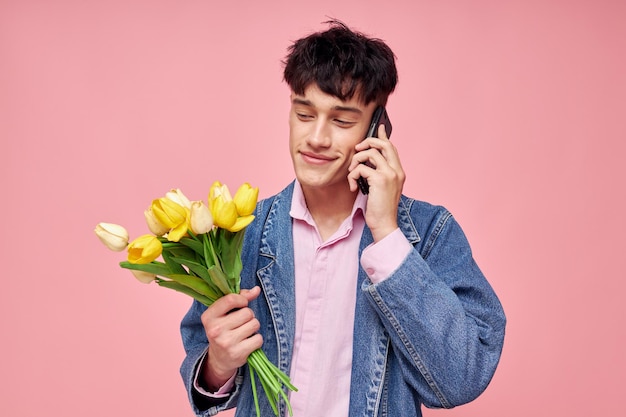 Young guy in a denim jacket with a bouquet of flowers talking on the phone pink background