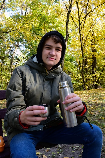 A young guy drinks tea from a thermos in an autumn park Dressed in a jacket with a hood