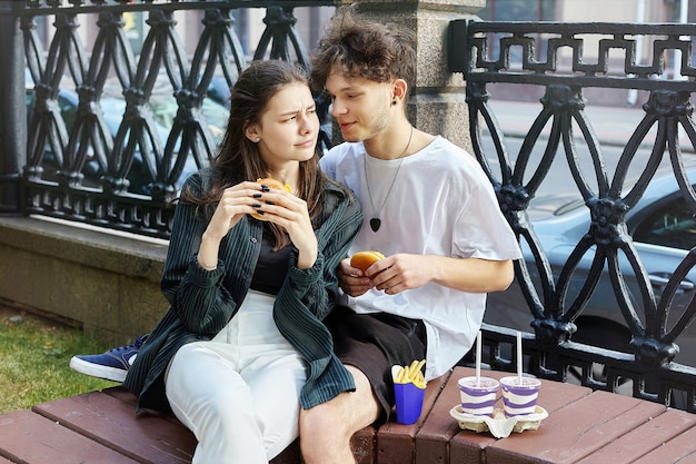 Young guy and a girl in the city are sitting on a bench and eating hamburgers..