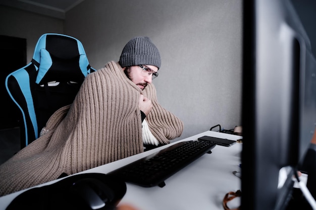 Young guy in glasses and a warm hat wrapped in a blanket works at a computer It39s cold in the room The concept of turning off heating and gas lowering the temperature in the room