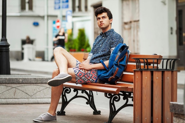 A young guy is sitting on a bench in the city Brunette in shorts and a blue shirt with a backpack